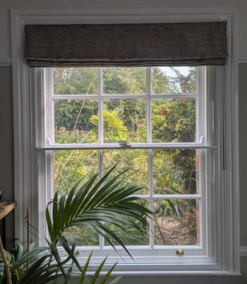 Heritage window with secondary glazing, showcasing a view of a lush garden, framed by a stylish window treatment and indoor plants, emphasizing bespoke solutions for period properties.